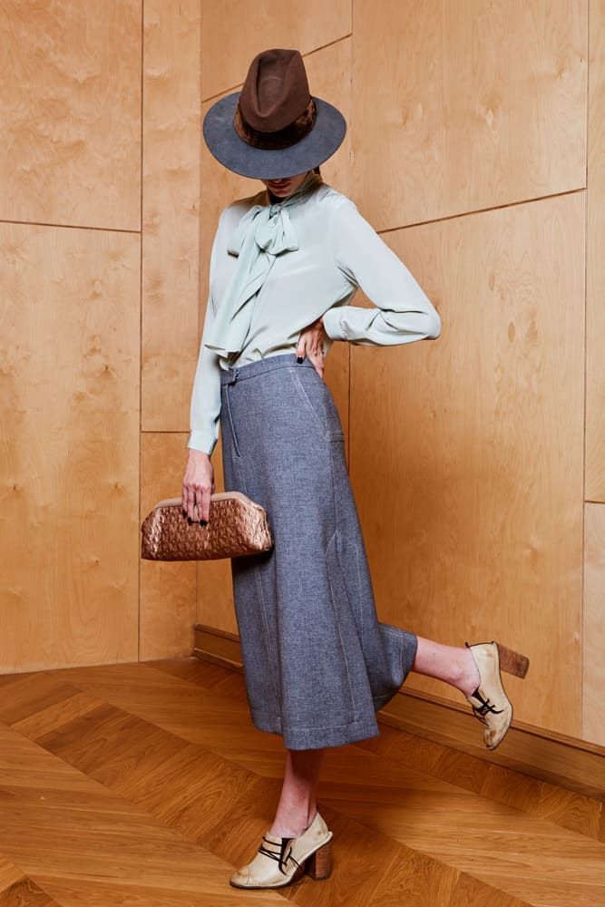 A woman wearing a long gray skirt, blouse and a fedora.